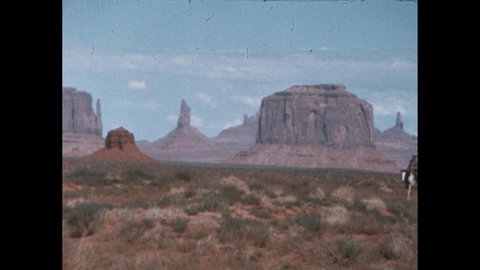 1880s: horseman leads horses through rocky desert landscape. Cowboy on horseback in remote landscape. Rock formations in American desert. Scrubland on hot day.