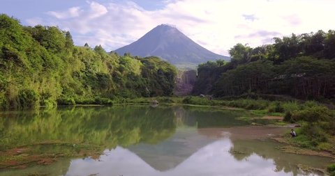 Aerial low altitude flight over tranquil nature lake surrounded by dense jungle and massive Merapi Volcano in background during sunlight