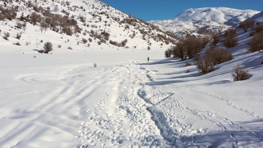 Man in khaki-colored hiking pants and black polo does Nordic walking in his mountain boots in a thick layer of snow in Mount Hermon in Israel on a sunny day. Aerial steady shot with vertigo affect