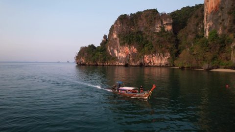 aerial drone pull away shot near Railay Beach in Krabi Thailand during sunrise as a Thai longtail boat motors through the Andaman Sea with a tourist onboard and large limestone mountains behind