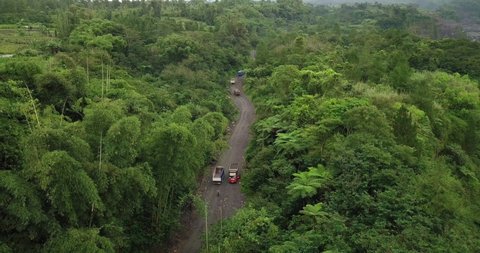 Aerial birds eye shot of trucks driving on rural road bet ween forest woodland on the slope of Merapi Volcano - Transport for sand mine in Indonesia - Ascend shot