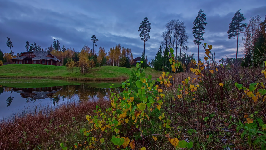 Static view of a wooden cottage in distance with dark clouds passing by in timelapse with lake surrounded by green grasslands in evening.