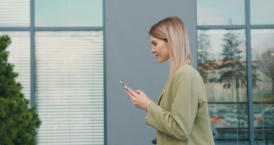 Side view of smiling young woman looking at mobile phone walking in the street to the office beside green trees pine. Outdoors Royalty-Free Stock Footage #1090460927