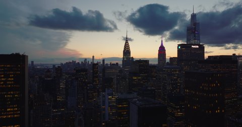 Forwards fly above city at twilight. Illuminated tops of iconic skyscrapers. Chrysler and Empire State Building against sunset sky. Manhattan, New York City, USA in 2021