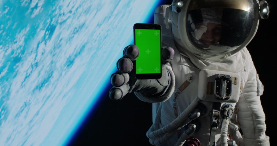 CHROMA KEY Caucasian female astronaut using her mobile phone during spacewalk near planet Earth, showing green screen into camera. Shot with 2x anamorphic lens Royalty-Free Stock Footage #1090470343