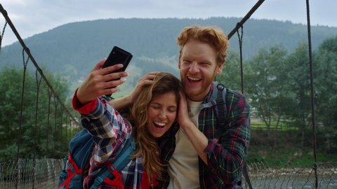 Friends taking selfie photo on smartphone. Tourists couple grimacing at mountains nature. Cheerful lovers kissing cheek on holiday trip. Smiling hipsters making video on travel blog. Leisure concept.
