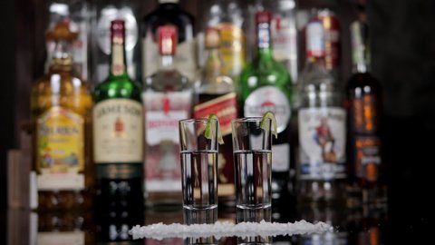 Стоковое видео: Close-up of a man and a woman taking shots of tequila with lime and salt in a bar. A couple drinks in a nightclub or bar against the background of shelves with bottles of alcohol.