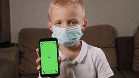 Portrait of a little boy in a medical mask at home on the couch, he holds a phone with a green screen.