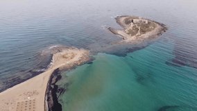 Aerial drone footage of the island and beach of Isola delle Correnti with an old lighthouse surrounded by clear turquoise sea water. Southernmost point in Sicily, Portopalo di Capo Passero, Italy.