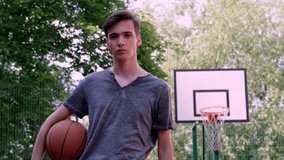 Teenager is going with a basketball in his hand is on the sport ground. Teenager on a basketball court goes to the camera with a basketball in his hands.   Confident face of a basketball player.