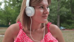 Portrait of a young woman in white large headphones in the park. Woman listening to music while sitting. slow motion