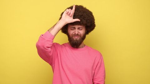 Bearded man with Afro hairstyle showing looser gesture holding fingers near forehead, sad because of silly mistake, wearing pink sweatshirt. Indoor studio shot isolated on yellow background.