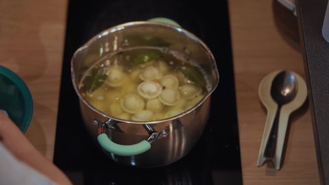 Person takes handful of dumplings in hands and throws them into boiling water in saucepan. Woman cooks pelmeni in aluminum pan on glass-ceramic kitchen stove. Cooking lunch or dinner for family.