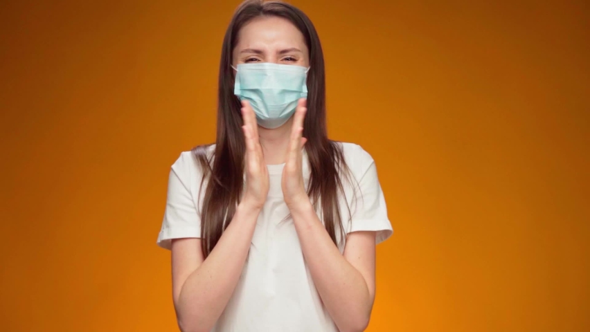 Woman in medical mask on face is very happy and claps her hands against yellow background