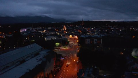 Forwards fly above street lit by orange street lights. Aerial view of town at dusk. Overcast sky. Killarney, Ireland