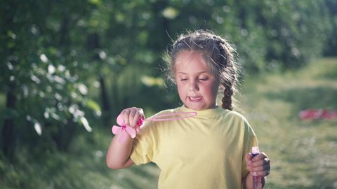 Happy little girl in park. Cheerful child blowing soap bubbles in nature. Forest park in summer. Dream and fun of girl on the green grass. Small soap bubbles are flying in wind. Happy child in park