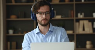 Young businessman in glasses wear headphones sit at office use laptop take part in online negotiation, lead business meeting via videocall app. Virtual meeting, tuition, on-line training event concept