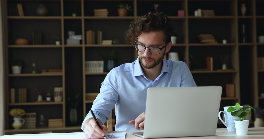 Young businessman employee in eyeglasses sit at desk in home office work use laptop, makes financial stats analysis, review online project revenue, take notes looks focused. Trading, workflow concept | Shutterstock HD Video #1090499419