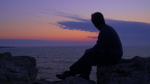 Young man sitting on a rock, watching the sea at dawn, waiting for the sunrise. Windy shore with dark blue water, dark blue clouds, cliff silhouettes and the Belt of Venus on the horizon. Middle shot.