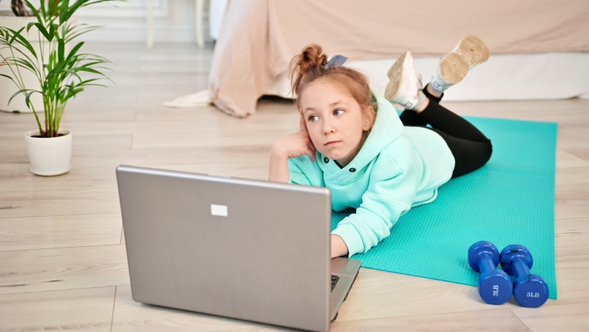 The girl is thinking and looking for sports exercises in the laptop. Baby heifer lying near laptop in the living room. Children and sports at home Royalty-Free Stock Footage #1090505989
