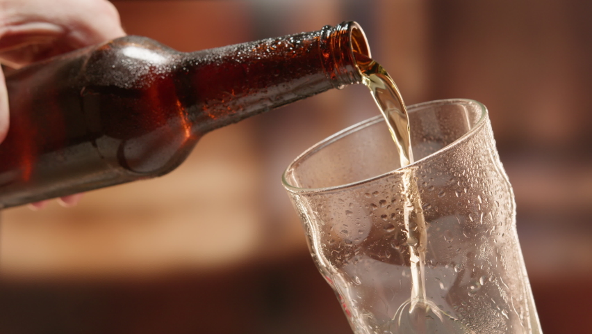 Beer is poured from dark brown bottle into beer glass in slow motion. Close-up light fresh beer poured into glass steamed up from cold. Lager beer foams and pours from bottle into glass. Royalty-Free Stock Footage #1090506995
