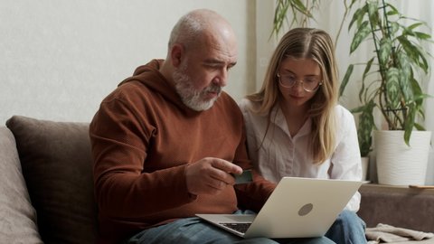 A pretty young woman shows her older mature father how to use a bank card and computer at home. An adult daughter teaches laptop apps to a focused middle-aged dad using modern technology.
