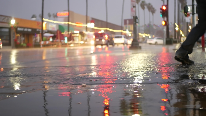 Cars lights reflection on road in rainy weather. Rain drops on wet asphalt of city street in USA, water raindrops falling on sidewalk. Palm trees and rainfall, twilight dusk. Ocean Beach, California.