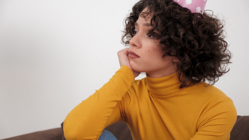 Close up upset curly woman in party hat looking away celebration holiday birthday alone at home. Sad brunette hispanic female wearing yellow turtleneck sitting on couch at home. Quarantine, lockdown