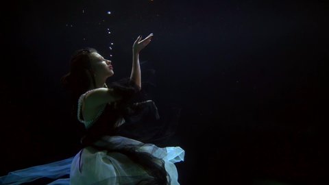 a woman in a magnificent dress hovers in the dark water and stretches hands to the surface. profile view