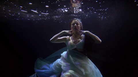 a brunette in a light dress hovers in dark water and moves hands beautifully in front of face. the middle plan