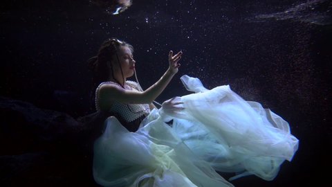 a woman in a lush light dress hovered in the dark water with bubbles and beautifully moves hands. profile view