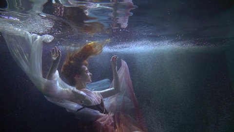 a red-haired woman in a pink and white dress and corset floats in dark water