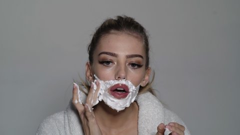 Attractive young woman shaves face. Cheerful funny woman has having cream over her face. She is shaving herself with razor.