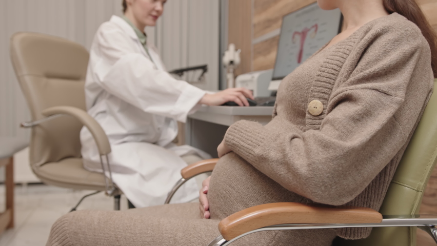 Slowmo of unrecognizable pregnant woman stroking her belly with tenderness while listening to her female obstetrician having appointment in clinic