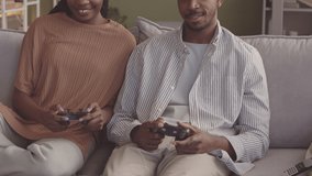 Slowmo of young cheerful African American couple using controllers while playing video games together, sitting on sofa in minimalist eco style living room