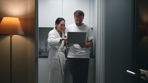 Diverse couple in sleepwear shop online on laptop standing together in kitchen. Asian wife and caucasian husband use computer at home