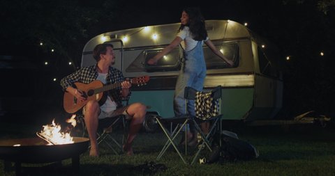 Cinematic shot of young husband is playing guitar and singing to his wife dancing carefree near their trailer with lighting while enjoying together romantic trip with camping caravan at night.