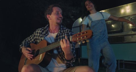 Cinematic shot of young husband is playing guitar and singing to his wife dancing carefree near their trailer with lighting while enjoying together romantic trip with camping caravan at night.
