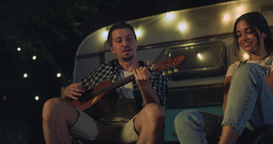Cinematic shot of happy carefree husband is playing guitar and singing to his wife both sitting near their trailer with lighting while enjoying together romantic trip with camping caravan at night. Royalty-Free Stock Footage #1090528983
