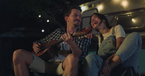 Cinematic shot of happy carefree husband is playing guitar and singing to his wife both sitting near their trailer with lighting while enjoying together romantic trip with camping caravan at night.