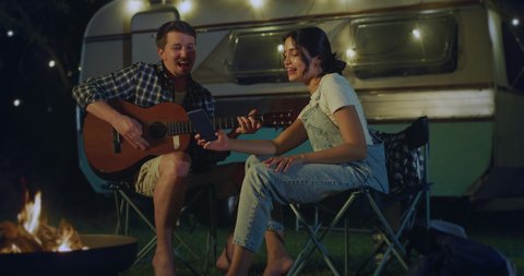 Cinematic shot of happy carefree wife is making video with smart phone while her husband is playing guitar and singing to her during their romantic trip with camping caravan at night.