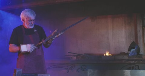 Side view of blacksmith with grey hair and beard holding long sword, inspecting. Old man wearing red apron and gloves, working in smithy, blue radiance around. Concept of forging.
