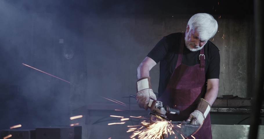 Front view of a blacksmith working hard in workshop. Old strong male with grey hair and beard, wearing apron, sharpening ironwork, sparks around. Concept of craftsmanship.