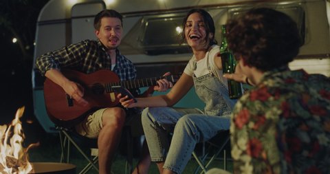 Cinematic shot of young happy carefree friends singing and dancing while one of them playing guitar during their relaxing vacation together with camping caravan at night.