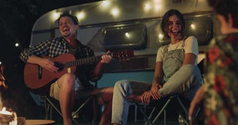Cinematic shot of young happy carefree friends singing and dancing while one of them playing guitar during their relaxing vacation together with camping caravan at night.