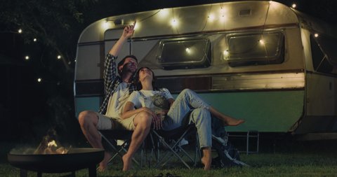 Cinematic shot of happy carefree romantic couple in love enjoys watching stargazing together at night starry sky near their trailer with lighting during romantic trip with camping caravan.