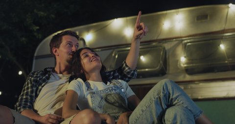 Cinematic close up shot of happy carefree romantic couple in love enjoys watching stargazing together at night starry sky near their trailer with lighting during romantic trip with camping caravan.