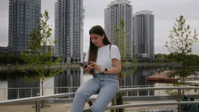 A young brunette girl sits in a park on a railing and communicates by video call on the phone. The girl is wearing a white T-shirt and jeans. Communication by phone, video communication, modern world