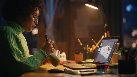 Black Female Artist Puts Unique Designs on Shoe Comparing with 3D Model on the Laptop Screen. Brasilian Woman Drawing at Evening. Hand Made and Craft Concept. Establishing Shot