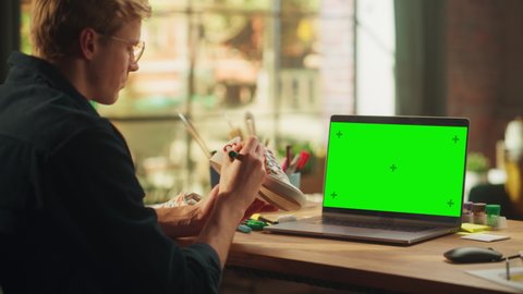 Male Designer Drawing on Shoe in front of Laptop Computer with Green Screen Mock Up Chroma Key. Teenage Freelancer Designing Unique Footwear at his Workshop for Online Sale Exclusive